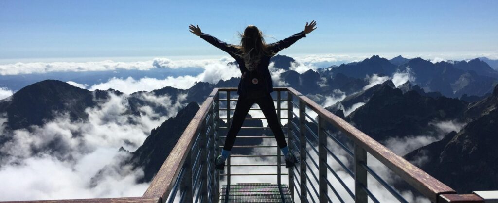A woman stands on a viewing platform in Vysoké Tatry, Slovakia, surrounded by clouds and mountains, embracing freedom.
