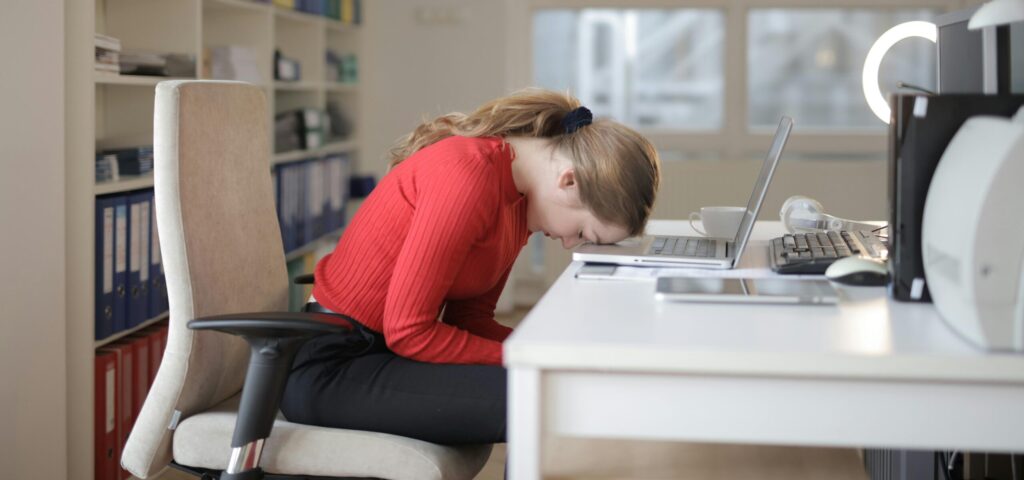 Štedrosť bez viny Tired woman in red sweater naps on office desk beside laptop, overwhelmed by remote work.